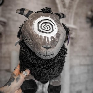 May include: A plush sheep toy with a grey face and a white spiral eye design. The sheep has black and striped horns, a black fluffy body, and black hooves. The toy is held up against a blurred background.