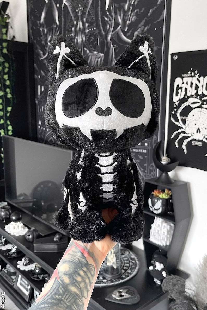 May include: Black plush cat skeleton toy with white bones. The cat has a white skull face with black eyes.