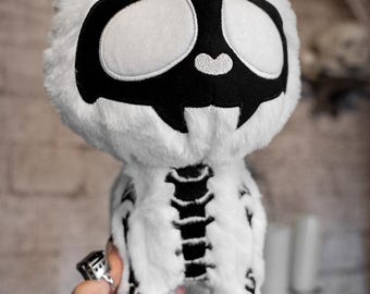 Op de afbeelding: Een witte pluche kat met een zwart skeletontwerp en een zwart schedelgezicht. De kat heeft een paars label op zijn oor. Het skelet van de kat is zichtbaar op zijn borst en poten.