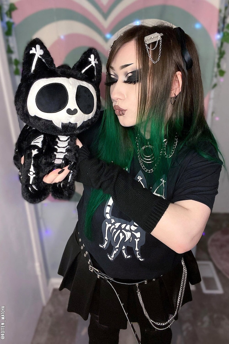 May include: A person with green and black hair is wearing a black t-shirt with a skeleton cat graphic, a black skirt, and black fingerless gloves. They are holding a black and white plush skeleton cat toy.