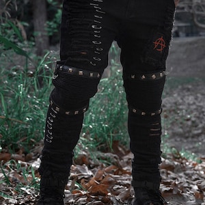 May include: Black distressed jeans with ripped details, metal accents, and a red embroidered symbol. The jeans feature studded leather bands around the knees and multiple safety pins along the side. A modern, edgy style.