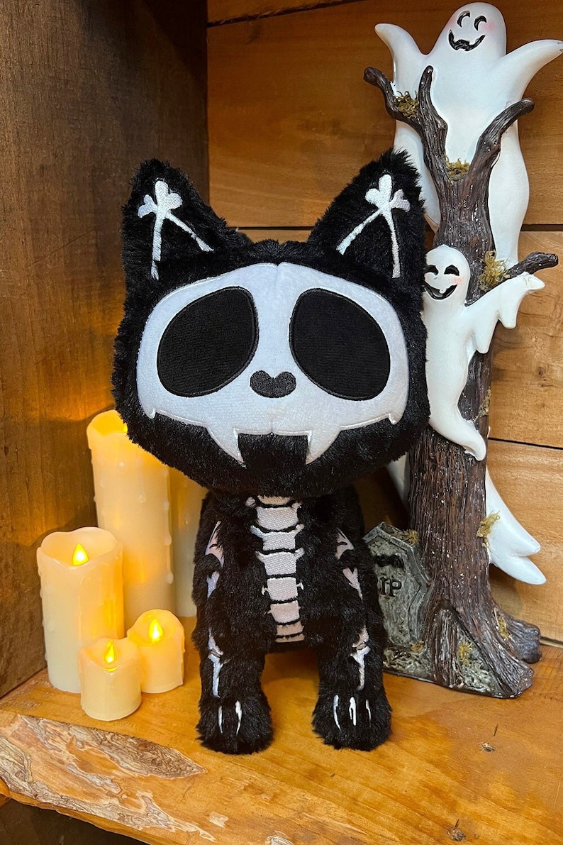 May include: Black plush skeleton cat with a white skull face and a black and white skeleton design on its body. The cat is sitting on a wooden shelf with candles and a ghost decoration.
