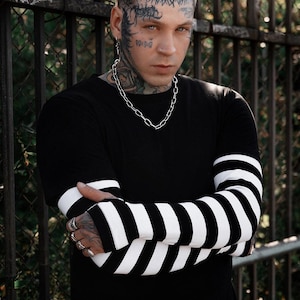 May include: A black long-sleeve shirt with white striped sleeves. The person in the image is wearing a silver chain necklace and multiple silver rings. The person has tattoos on their face and neck.