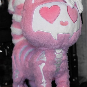 May include: A pink plush cat with a skull-shaped face and heart-shaped eyes. The cat has a white and pink striped tail and silver-colored skeleton details. The plush toy is a unique and whimsical design.