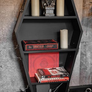 May include: Black coffin-shaped bookshelf with multiple shelves. The shelves hold candles, a gargoyle statue, and books, including one titled "Dracula." The bookshelf has decorative handles and is set against a brick wall.