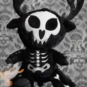 May include: A black plush toy in the shape of a deer with a skull face and antlers. The toy has a white skeleton design on its chest and legs. The toy is held by a person with a tattoo and gold jewelry.