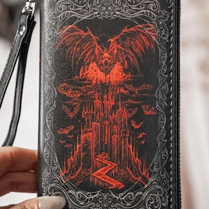 May include: Black wallet with a red gothic castle and bat design. The wallet has a wrist strap and a zipper closure. The artwork features a castle, bats, and a winged creature. The design is framed with an ornate border.