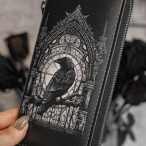 May include: Black wallet featuring a raven design. The wallet showcases a detailed illustration of a raven perched on a branch within a gothic-style stained-glass window. It has a zip closure and is held by a person.