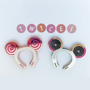 May include: Two gold and silver mouse ear headbands with the word "TWICE" and "ONCE" on them. The ears have a pink and black swirl design with the word "TWICE" in a circle.