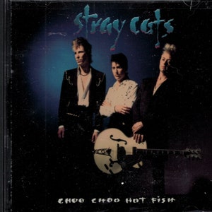 May include: A CD cover featuring a band called "Stray Cats" with the album title "Choo Choo Hot Fish". The band members are dressed in black leather jackets and jeans, and they are holding a guitar. The background is a dark blue and purple gradient.