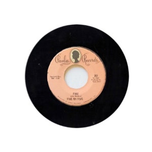 Może przedstawiać: Czarna płyta winylowa z różową etykietą. Etykieta zawiera tekst "Paula Records" i "Fire (Jim Hendrix) Five By Five". Numer płyty to 302.