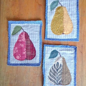 May include: Three rectangular fabric wall hangings featuring pear appliqués. Each piece has a white grid-patterned centre, a blue denim border, and a green leaf. The pears are made of multi-textured fabric patches in pink, yellow, and brown tones.