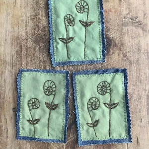 May include: Three square fabric patches with a light green background and a blue denim border. Each patch features an embroidered design of two stylized fern-like plants in brown thread. The patches are arranged on a wooden surface.