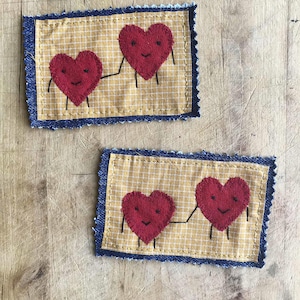 May include: Two rectangular patches with a yellow grid pattern and a blue denim border. Each patch features two red heart-shaped figures with smiling faces and black stick arms and legs, holding hands.