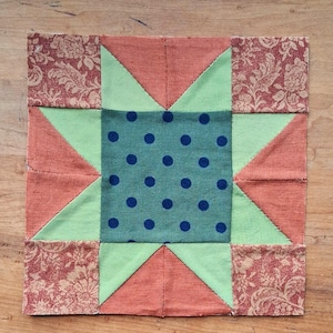 May include: A square quilt block featuring a central teal square with dark blue polka dots, surrounded by a star pattern made of light green triangles, solid orange triangles, and four corner squares with a reddish-brown floral pattern.