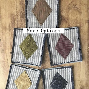 Peut inclure: Cinq dessous de verre en tissu avec des losanges de différentes couleurs : marron, vert olive, bordeaux, or et bleu marine. Chaque dessous de verre a un fond rayé et un bord effiloché. Le texte "More Options" est visible.