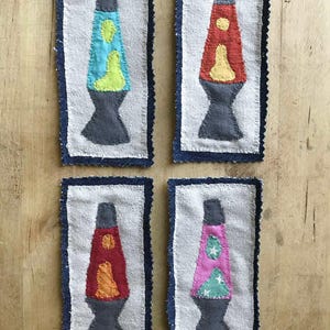 May include: Four handmade fabric patches with lava lamp designs. Each patch has a white background and a blue denim border. The lava lamps are in various colours, including blue, red, and pink, with contrasting lava-like shapes.