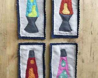 May include: Four handmade fabric patches featuring lava lamp designs. Each patch has a white background with a blue denim border. The lava lamps are in various colors, including blue, red, and pink, with contrasting lava-like shapes.