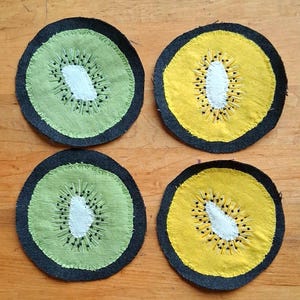 May include: Four circular fabric coasters featuring embroidered fruit slice designs. Two coasters are light green and two are yellow, each with a white center, radiating black embroidered seed details, and a dark charcoal gray fabric border.