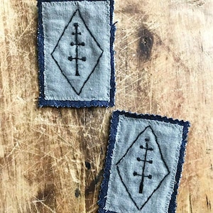 Peut inclure: Deux écussons rectangulaires avec un centre gris et une bordure bleu foncé. Chaque écusson présente un losange noir avec un motif d'arbre stylisé à l'intérieur. Les écussons sont placés sur une surface en bois.