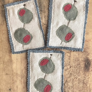 May include: Three rectangular fabric coasters featuring an appliqué design of two green olives with red pimentos on a cocktail stick. Each coaster has a cream-coloured centre panel with visible stitching and a frayed blue denim border trim.