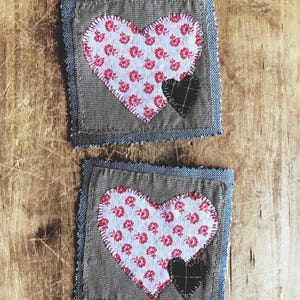 May include: Two square fabric coasters featuring a brown and white checkered base with frayed denim edges. Each coaster displays a large white heart with a red floral pattern and a smaller, solid black heart stitched onto the bottom right corner.