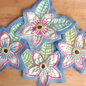 May include: Four fabric patches featuring white floral shapes embroidered with pink, blue, and yellow thread on light blue denim backgrounds. Each flower is surrounded by green embroidered leaves with visible stitching details on the petals.