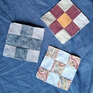 May include: Three square fabric coasters featuring patchwork designs. One coaster has a blue and white grid pattern, another features a central yellow square surrounded by red plaid and light floral squares, and the third displays paisley and blue fabric.