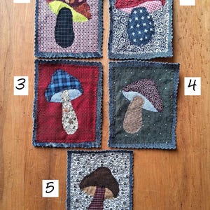 Peut inclure: Cinq écussons rectangulaires en tissu avec des appliqués de champignons. Le numéro 1 a un fond à motifs violets. Le 2 présente un champignon rouge sur fond floral. Le 3 montre un champignon à carreaux bleus sur fond rouge. Le 4 a un fond vert foncé. Le 5 affiche un champignon marron sur un imprimé floral blanc.