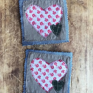 May include: Two handmade fabric heart coasters with a denim border. The hearts are made of a white fabric with a red floral pattern and a smaller green heart in the centre.