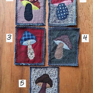 May include: Five rectangular fabric patches featuring mushroom appliqués. Patch 1 has a patterned mushroom on a geometric background. Patch 2 shows a red-capped mushroom on a floral background. Patch 3 features a blue-capped mushroom on red fabric. Patch 4 displays a dark mushroom on a dotted background. Patch 5 shows a brown mushroom on a floral print.