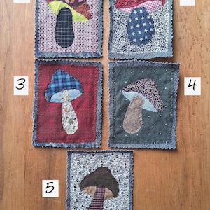 May include: Five rectangular fabric patches featuring appliquéd mushroom designs. Each patch has a frayed denim border and displays a unique combination of patterned textiles, including floral, plaid, and polka dot fabrics in various colors.