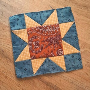 May include: A square fabric quilt block featuring a central orange square with a dark brown paisley pattern. The center is surrounded by a yellow and white checkered star shape, set against a dark blue background with a small black floral print.