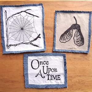 May include: Three square fabric patches with frayed denim borders. One patch features a black ink spiderweb illustration, another shows a black ink maple seed pod, and the third displays the black text "Once Upon A Time" in a serif font.