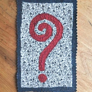 May include: A rectangular fabric patch featuring a white background with a dense black floral pattern. A large, textured red question mark is appliqued in the center. The patch has a dark blue denim border with a frayed, zig-zag stitched edge.