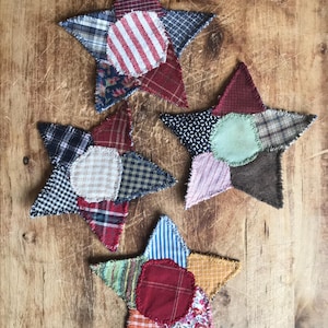 May include: Three handmade patchwork stars featuring a variety of fabrics in red, blue, green, brown, and white. The stars are made from different patterns of plaid, striped, and floral fabrics.
