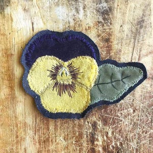 May include: A fabric patch shaped like a pansy flower, featuring a dark purple upper petal, a bright yellow lower petal with dark brown stitched details, and a single olive green leaf with black embroidered veins, all outlined in dark blue denim.