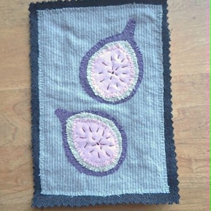 May include: A rectangular fabric patch featuring a light blue pinstriped background with two appliqued fig halves. The figs have purple outer skins, light green rings, and pale pink centers with dark stitched details, bordered by a black zigzag edge.