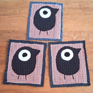 May include: Three rectangular fabric coasters featuring a black bird appliqué with a white eye on a red and white chequered background. Each coaster is bordered by a dark blue denim-like fabric with frayed, zigzag-cut edges on all four sides.