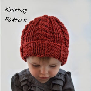 Puede incluir: Un niño lleva un gorro de punto rojo con un patrón de trenza y una camisa gris con volantes y sostiene una piña.