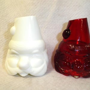 Puede incluir: Dos recipientes de vidrio con forma de Papá Noel. Uno es blanco, el otro rojo. Ambos tienen una parte superior cónica y un diseño de rostro detallado. El blanco tiene un acabado liso, mientras que el rojo es translúcido.