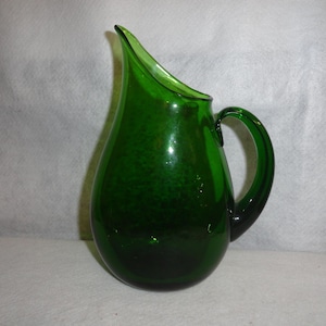 May include: A green glass pitcher with a teardrop shape and a handle. The pitcher has a textured surface.