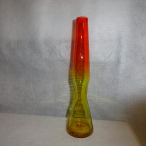 May include: A tall, slender glass vase with a vibrant color gradient. The top is a bright red, transitioning to orange, and then to a yellow base. The vase has a unique, hourglass-like shape.