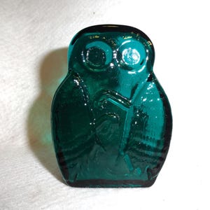 May include: A teal-colored glass owl figurine. The owl has large, round eyes and a detailed depiction of feathers. The figurine is a solid, decorative piece, likely for display. The glass has a glossy finish.