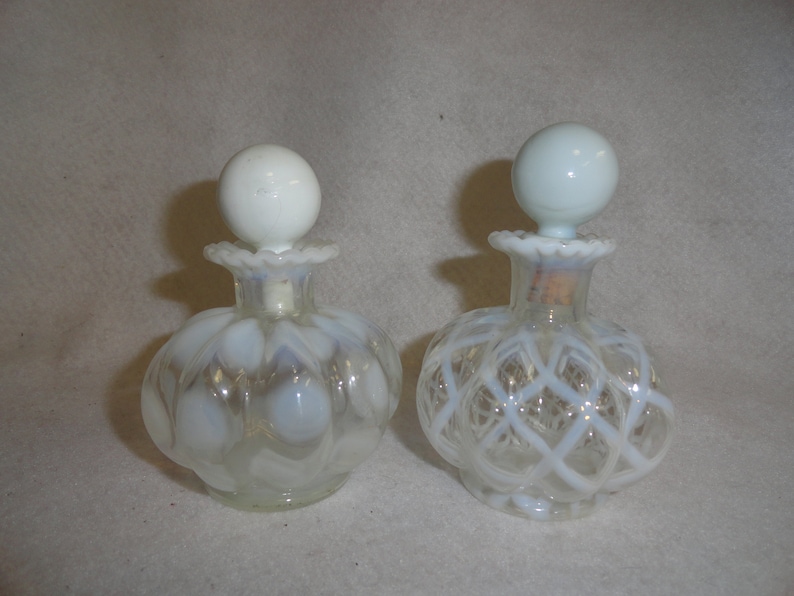 May include: Two clear glass perfume bottles with opalescent white glass stoppers. The left bottle features a rounded, ribbed body, while the right bottle displays a raised white lattice pattern. Both have ruffled rims and spherical tops.