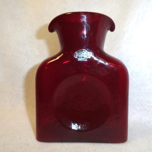 May include: A deep red glass vase with a wide, flared rim and a flat, rectangular body. The front features a circular indentation in the center and a small white sticker near the neck that reads Viking Glass Made in USA in black text.