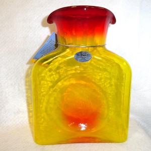 May include: A Blenko handcrafted glass vase with a square body and a flared top. The vase features a gradient of colors, transitioning from red at the top to yellow and orange in the center. A silver wire is wrapped around the neck.