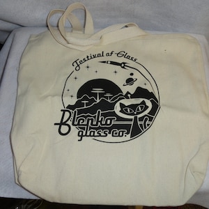 May include: A white canvas tote bag with black print. The print features a cat in a space helmet, a rocket ship, and the text "Festival of Glass Blenko glass co."