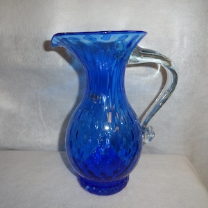 May include: A cobalt blue glass pitcher with a clear glass handle. The pitcher has a textured, bubbled surface and a flared lip.