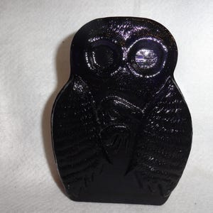 May include: A dark, glossy, owl-shaped decorative item. The owl has large, round eyes and detailed wing and feather patterns. The item is a solid, dark color, with a smooth, reflective surface.